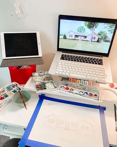 Workspace with laptop, tablet, watercolor paints, and house illustration in progress.