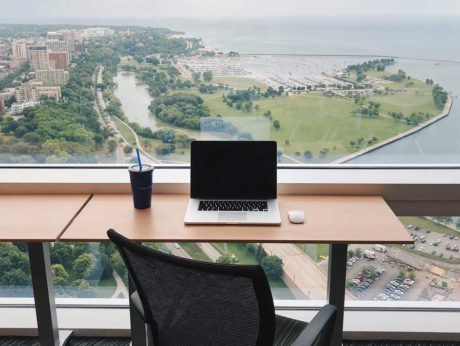 Laptop on a wooden desk with a view of a city park and waterfront through a large window.