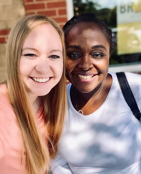 Two women smiling outdoors in natural light in front of a brick building.