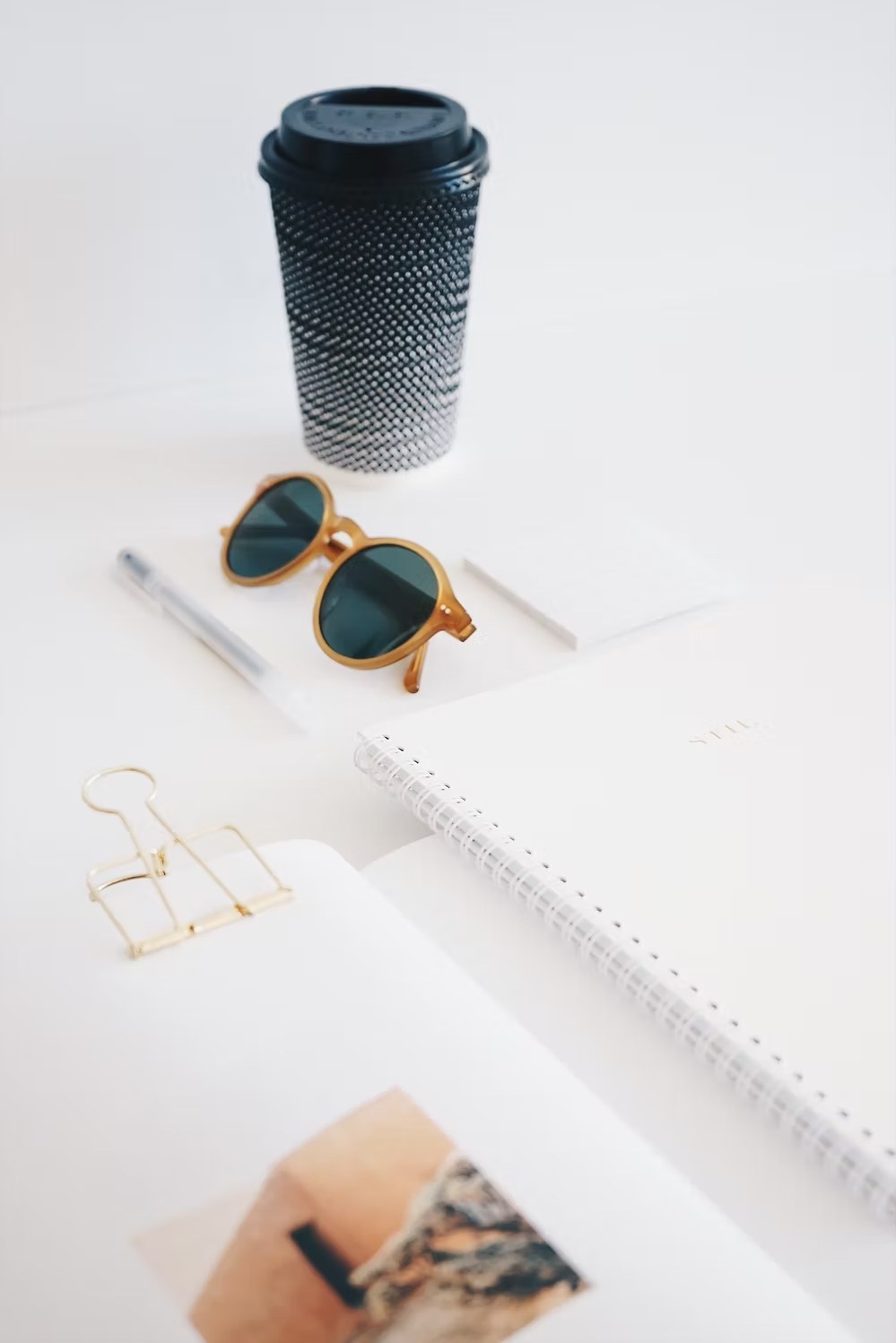 Minimalist desktop scene with sunglasses, coffee cup, notebook, and gold binder clip on white surface.