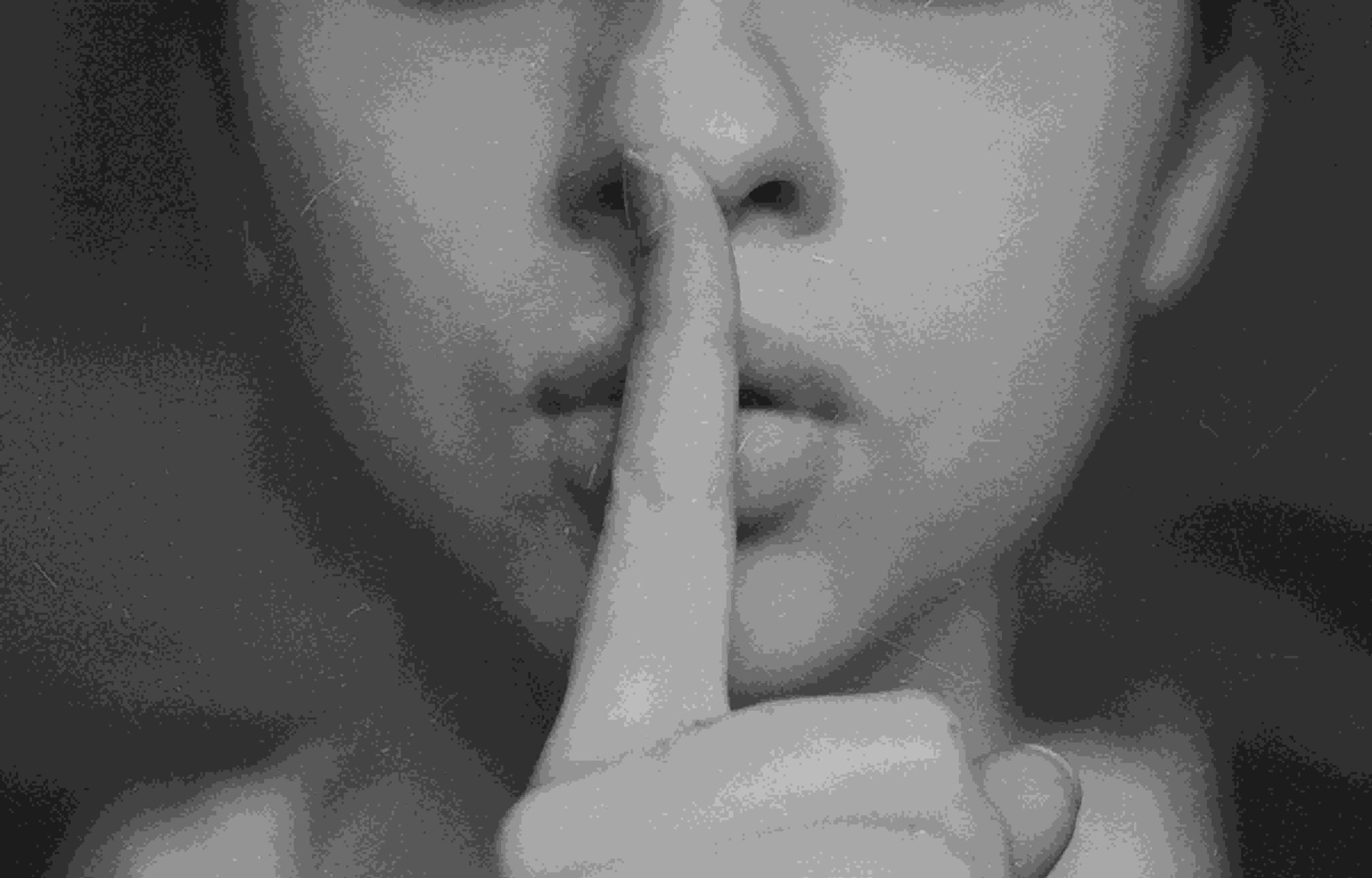 Close-up black-and-white image of a person making a shushing gesture with a finger over their lips.