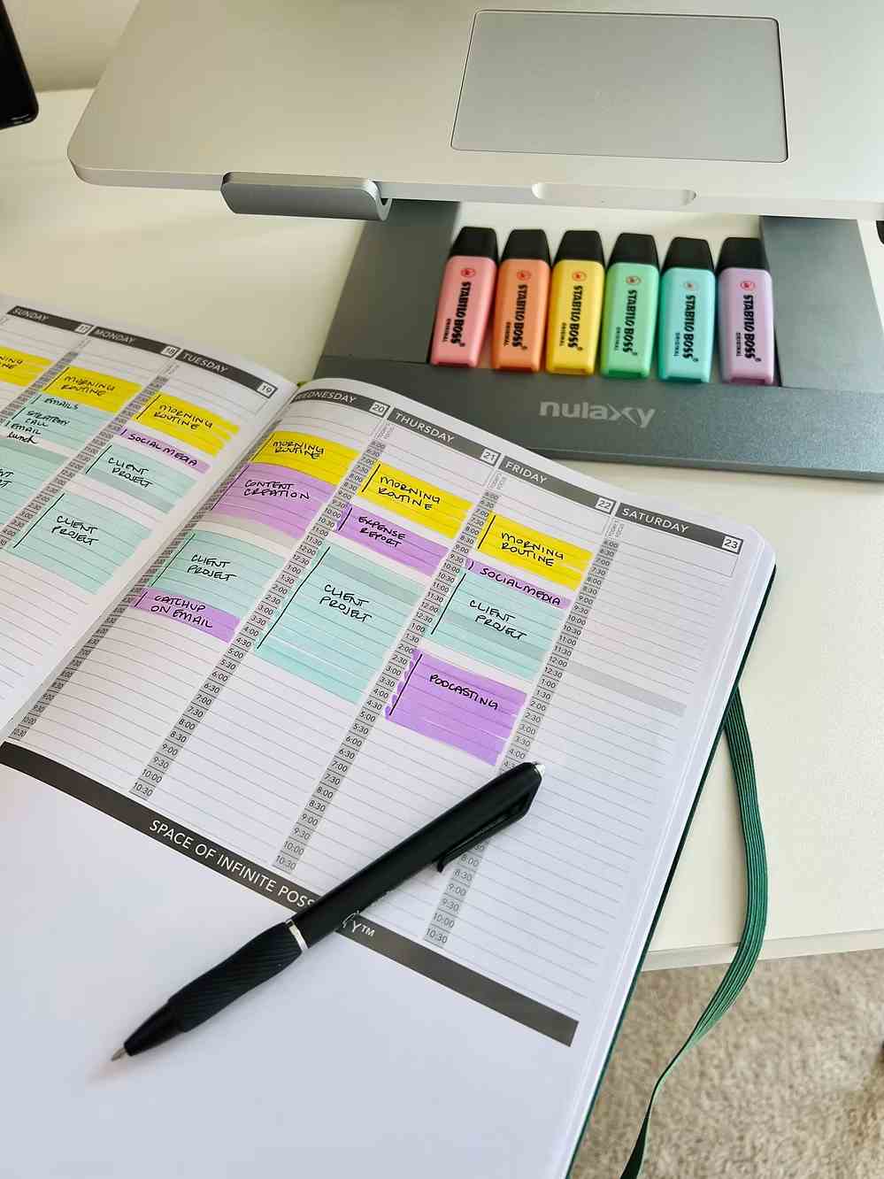 Open planner on desk with black pen and colorful highlighters beneath a laptop stand.