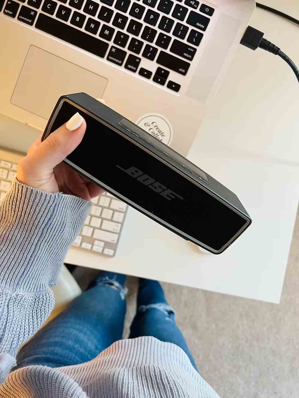 Person holding a Bose Bluetooth speaker above a laptop and keyboard on a white workspace.