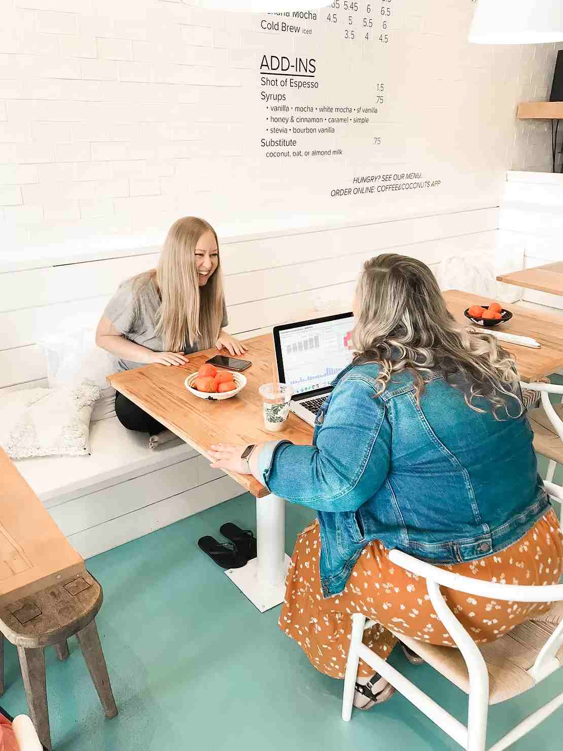 Two women meeting in a bright café, laughing and working together at a laptop.