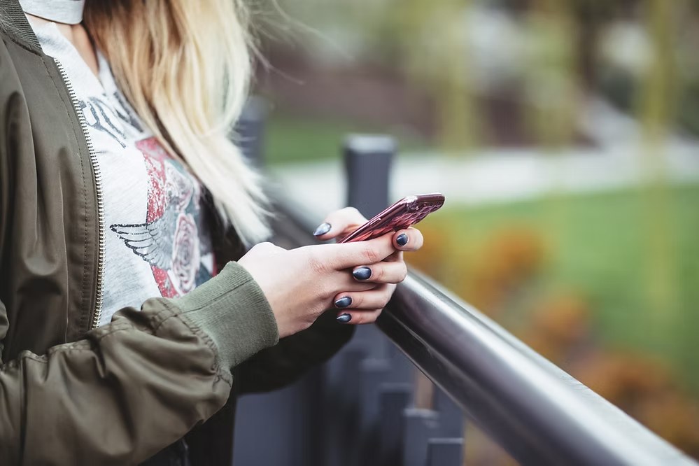 Close-up of woman using a smartphone with dark nail polish and graphic T-shirt outdoors.