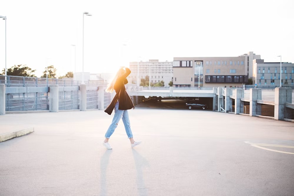 Woman walking across rooftop parking lot in casual outfit with city buildings in the background.