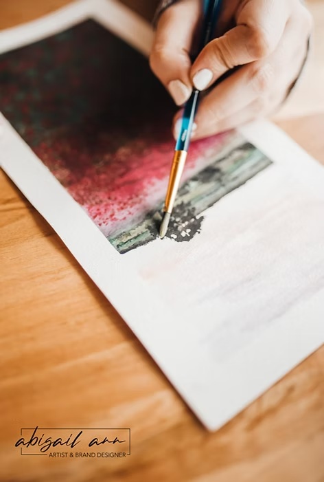 Hand painting a colorful abstract design on watercolor paper with a fine brush on a wooden desk.