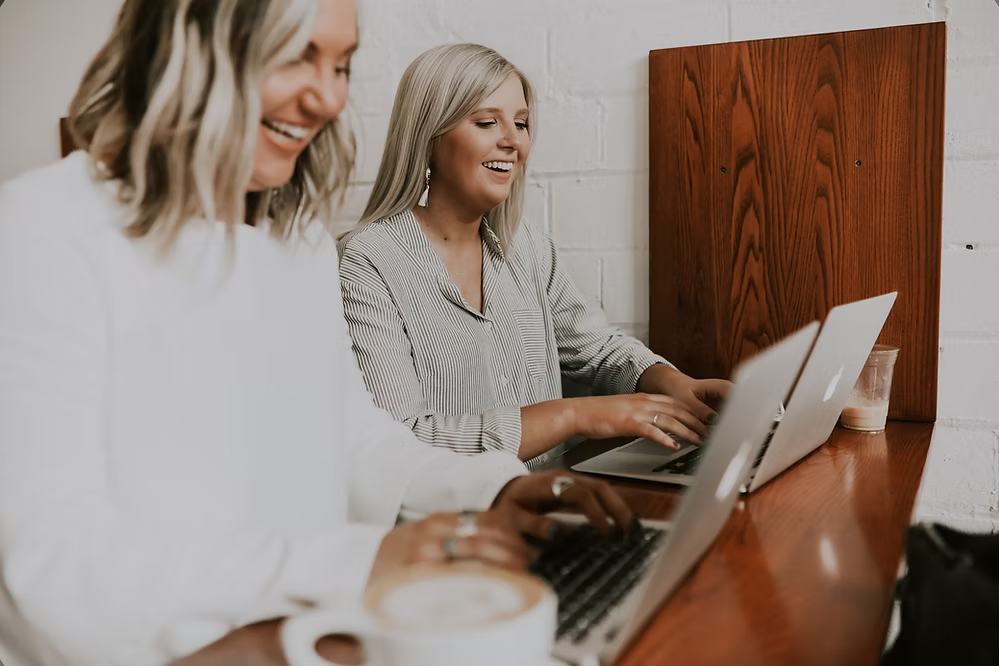 Two women smiling and working on laptops together at a cafe wooden counter.