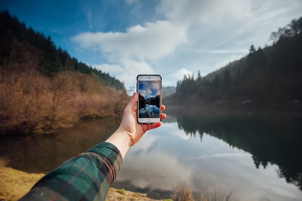 Hand holding a smartphone capturing a scenic lake reflection surrounded by trees and hills.