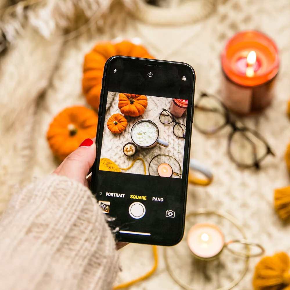 Person taking a cozy autumn-themed flat lay photo with pumpkins, candles, and a mug on an iPhone.