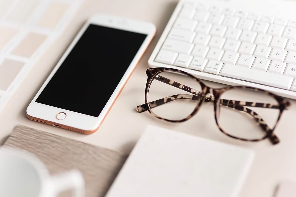 Close-up of smartphone, tortoiseshell eyeglasses, and white keyboard on a clean beige desk.