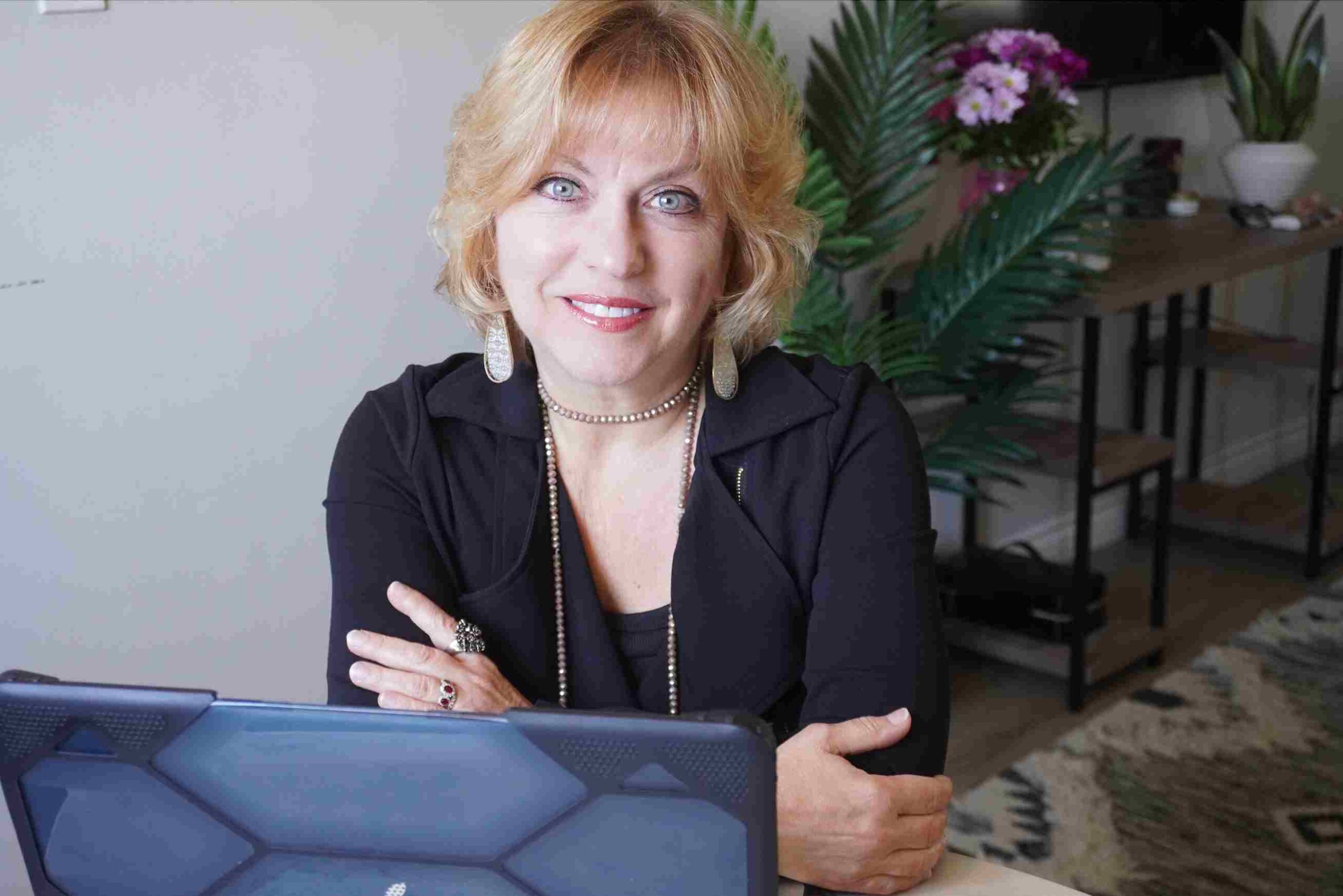 Woman with blonde hair wearing black, sitting confidently at a desk with a laptop.