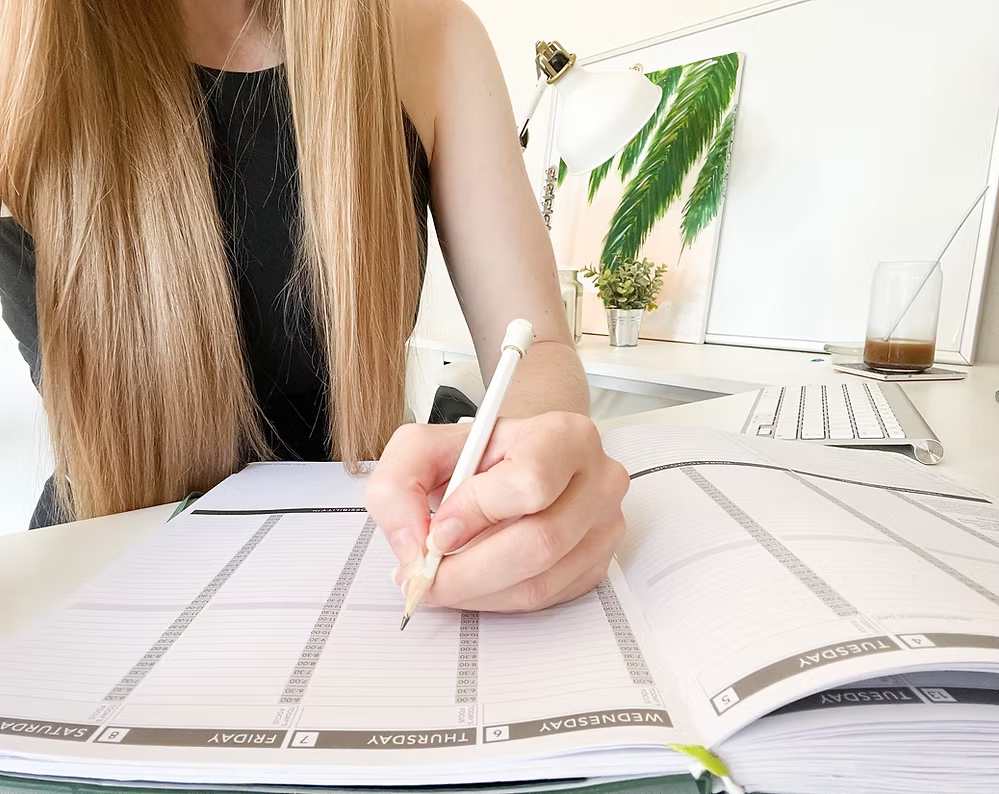 Close-up of woman's hands filling out a weekly calendar planner with pen at organized desk workspace.
