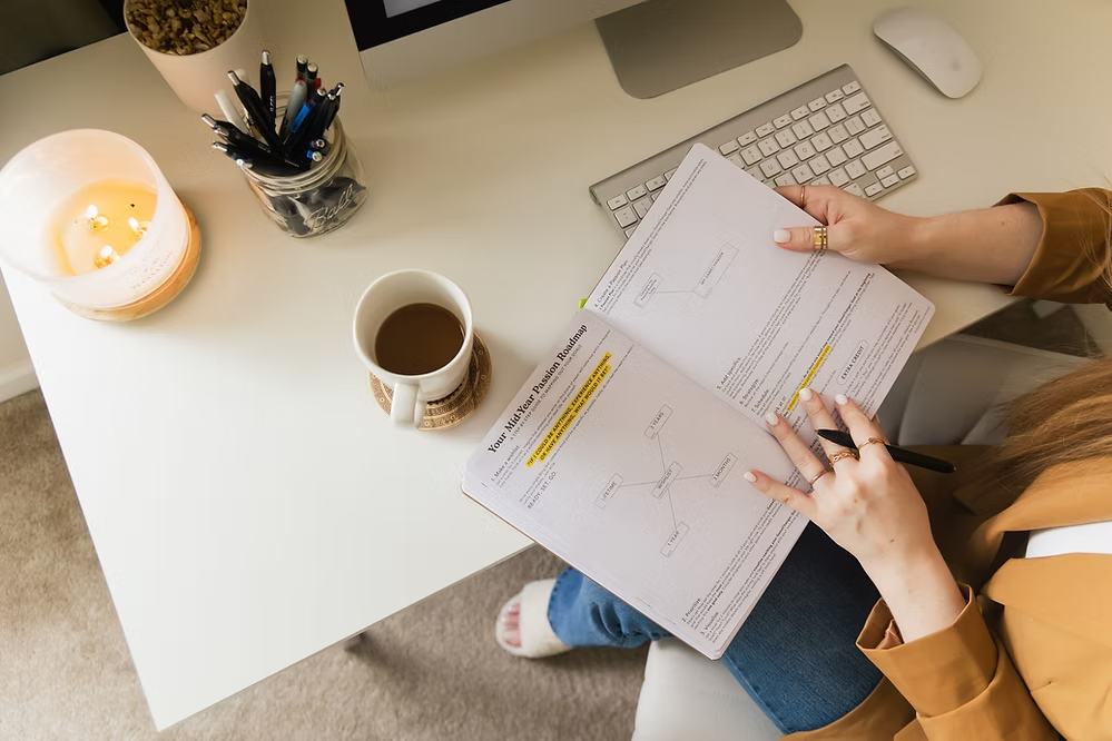Overhead view of woman reviewing documents and highlighting important sections at white desk with coffee and office supplies.