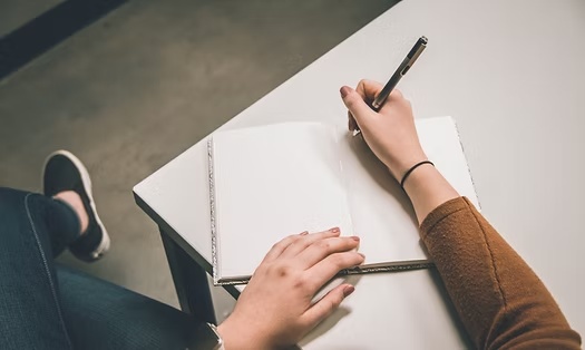 Person writing in an open notebook with a pen at a clean workspace.
