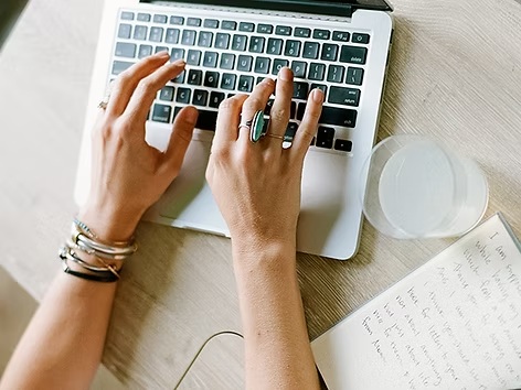 Hands typing on laptop keyboard with coffee cup and papers nearby in natural lighting.
