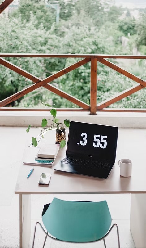 Minimalist workspace with laptop displaying time, small plant, and modern chair by large window with garden view.
