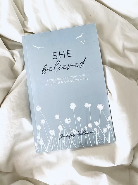 Light blue book titled "She Believed" by Jennifer Williams resting on white fabric background.