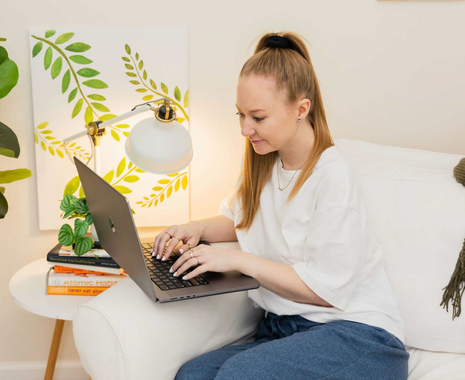 Woman typing on a laptop while sitting on a white couch surrounded by books and greenery.