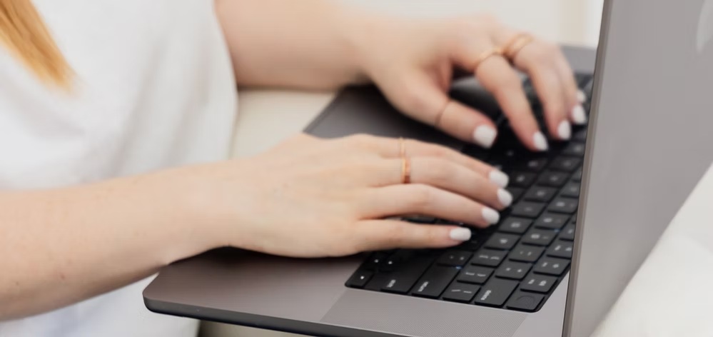 Woman typing on a laptop while sitting on a white couch surrounded by books and greenery.