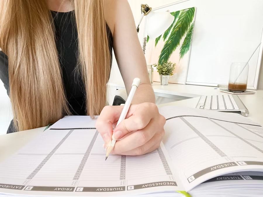 Woman writing in weekly planner with pencil at organized desk workspace with computer and plants visible.
