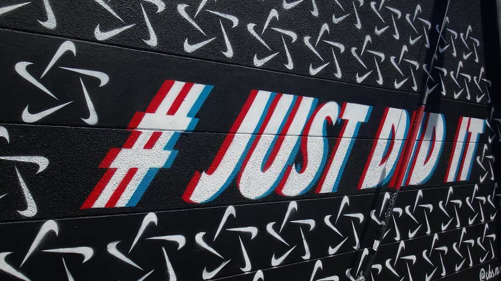 Nike "Just Do It" hashtag spray-painted on black wall with repeated Nike swoosh logos.