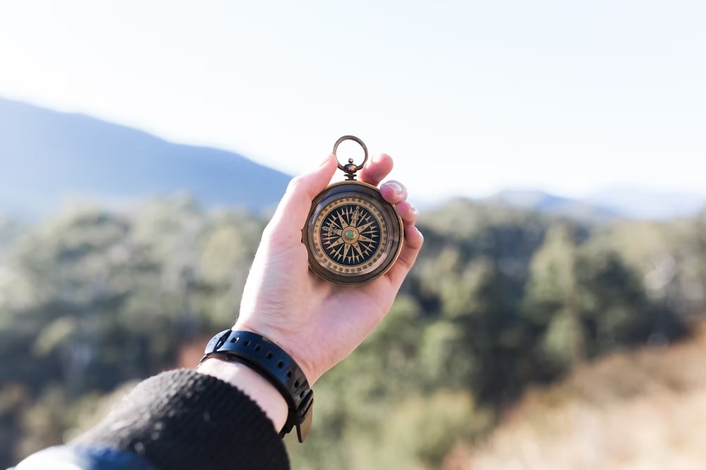 Hand holding vintage brass compass with mountain landscape in blurred background.