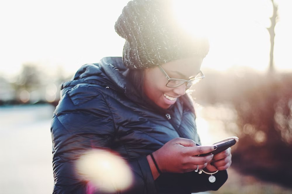 Smiling man in winter coat and beanie using smartphone outdoors with warm sunlight.