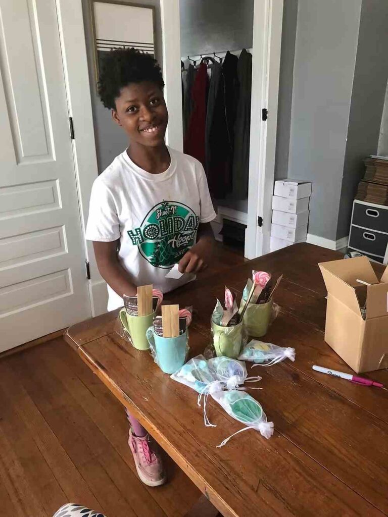 Young person sitting at wooden table packaging handmade products in green mugs with shipping materials.