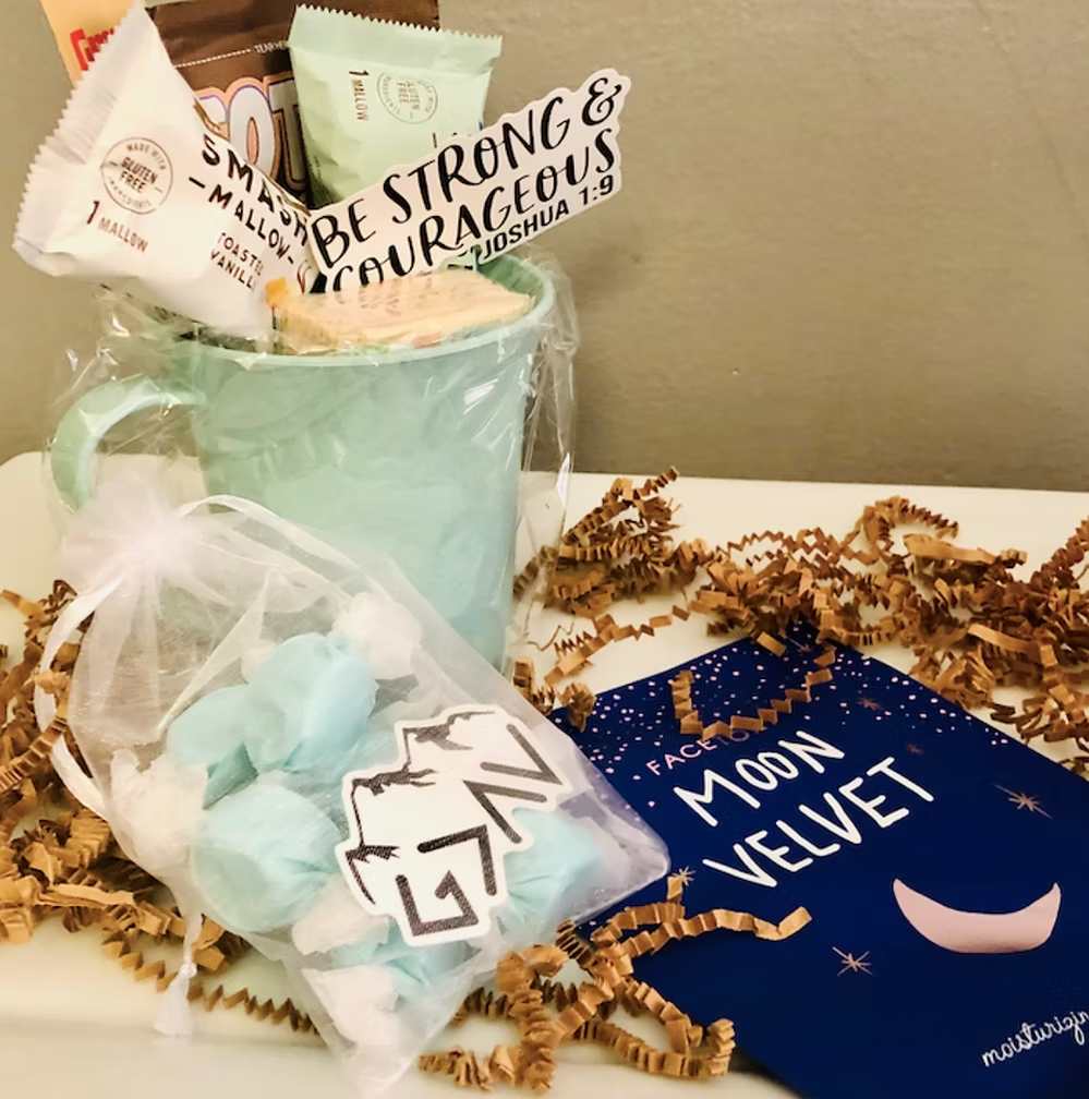 Gift basket with mint green mug, inspirational items, face mask, and "Be Strong & Courageous" card with brown paper filler.