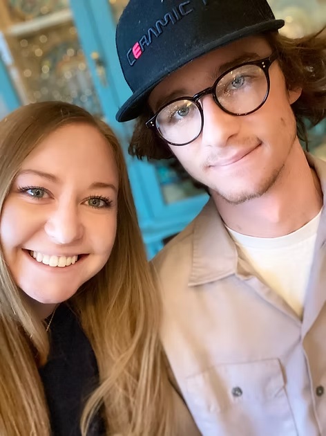 Happy young couple taking selfie together indoors, woman with long blonde hair and man wearing glasses and cap.