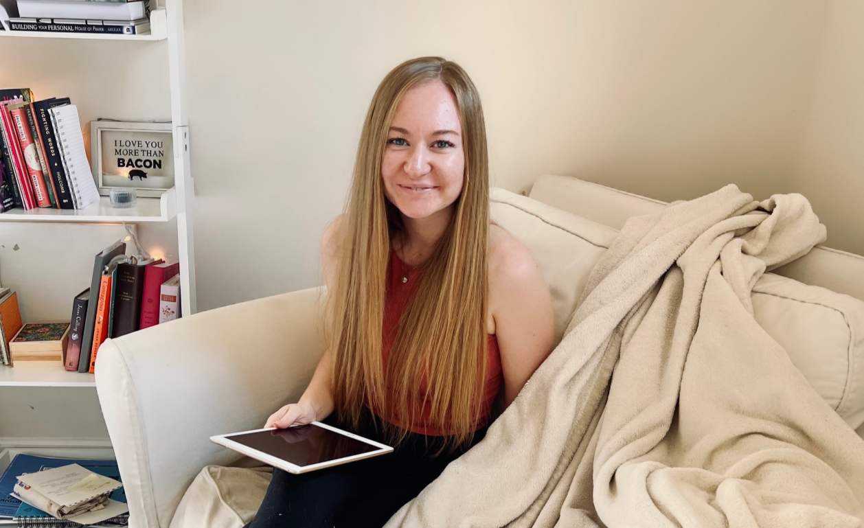Woman sitting on a couch with a tablet, surrounded by books and cozy blankets.