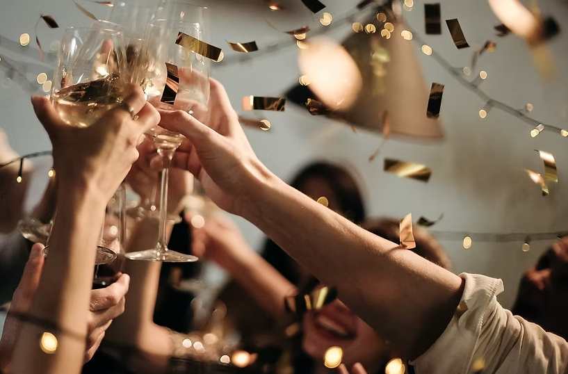 Group of people raising wine glasses in a celebratory toast with confetti and string lights 