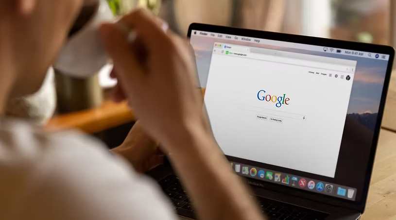 Person sitting at desk with coffee cup, laptop open to Google homepage 