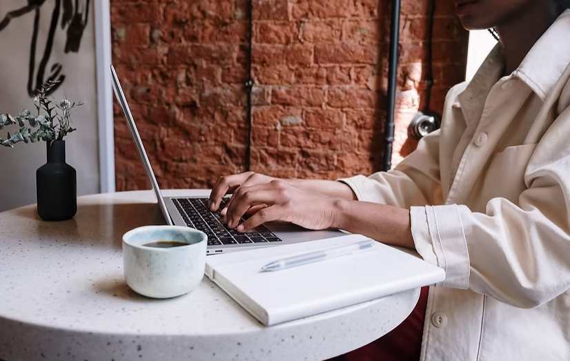 Person typing on a laptop at a round white table with notebook, pen, and coffee cup 