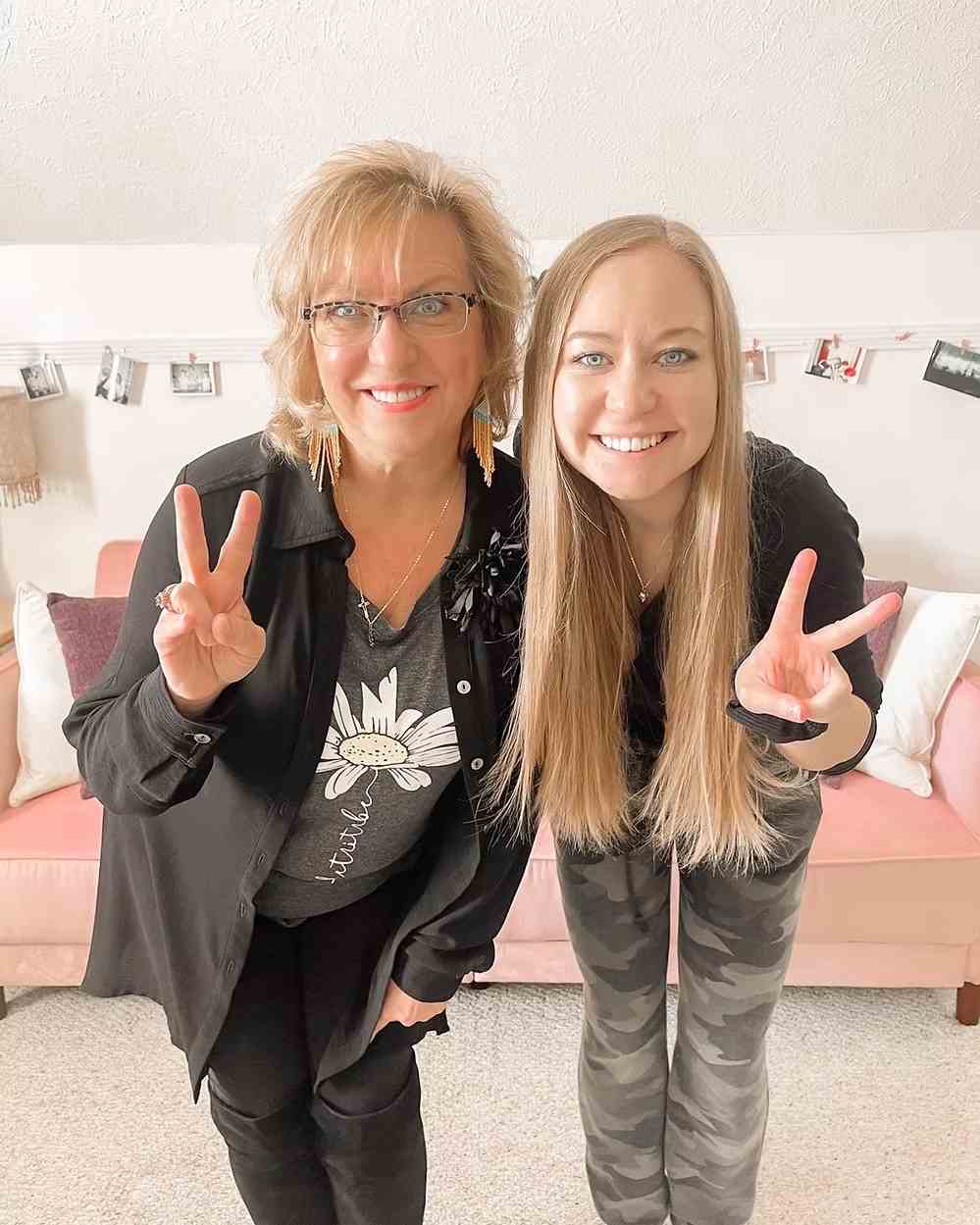 Two people smiling indoors, making peace signs in front of a pink couch with photo string decor.